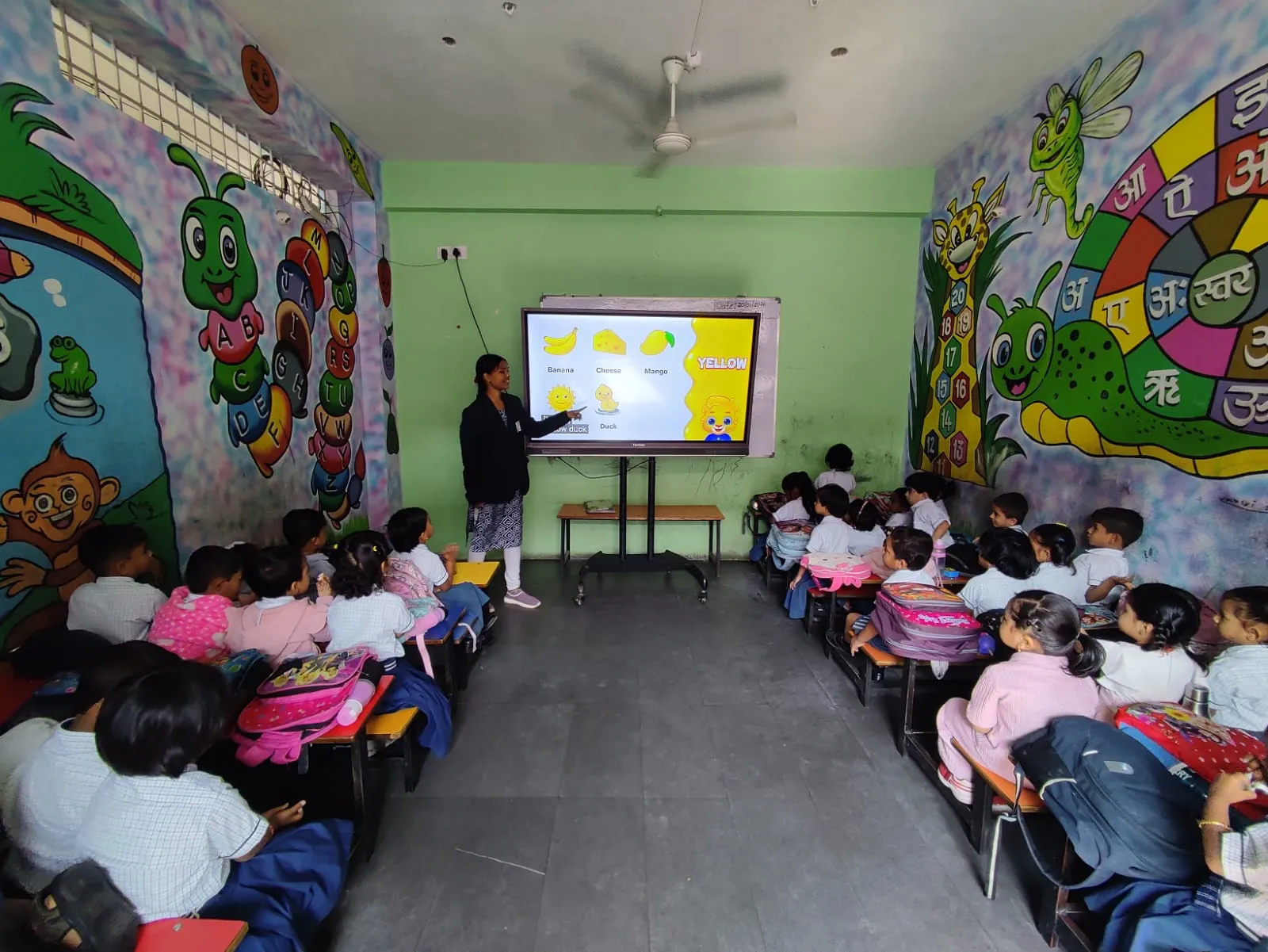 Teacher pointing at digital screen showing fruits while small children sit on benches in colorful classroom with cartoon wall art