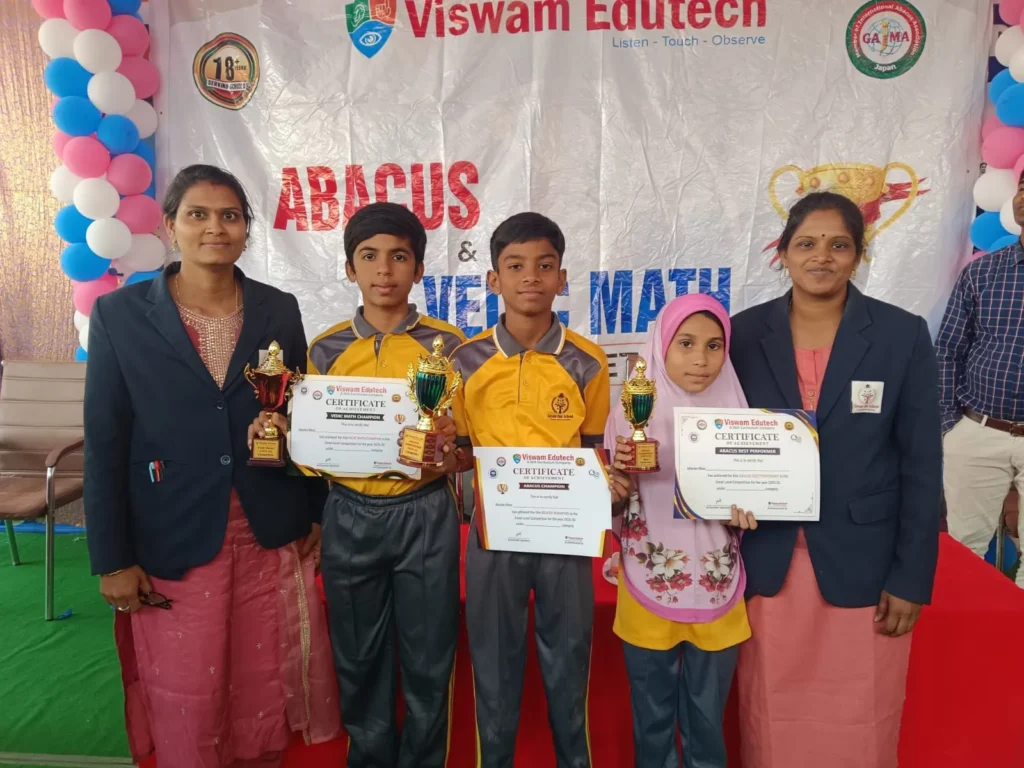 Students holding trophies and certificates with teachers on stage in front of an event banner during an achievement and award ceremony at Gnyan Oak School.