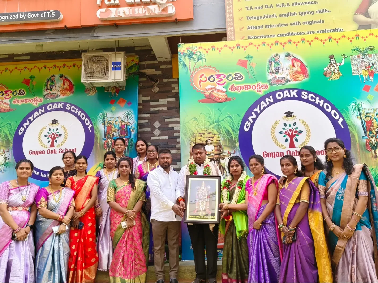 Group of staff members standing outside the school building holding a framed picture and smiling together as a dedicated faculty team.
