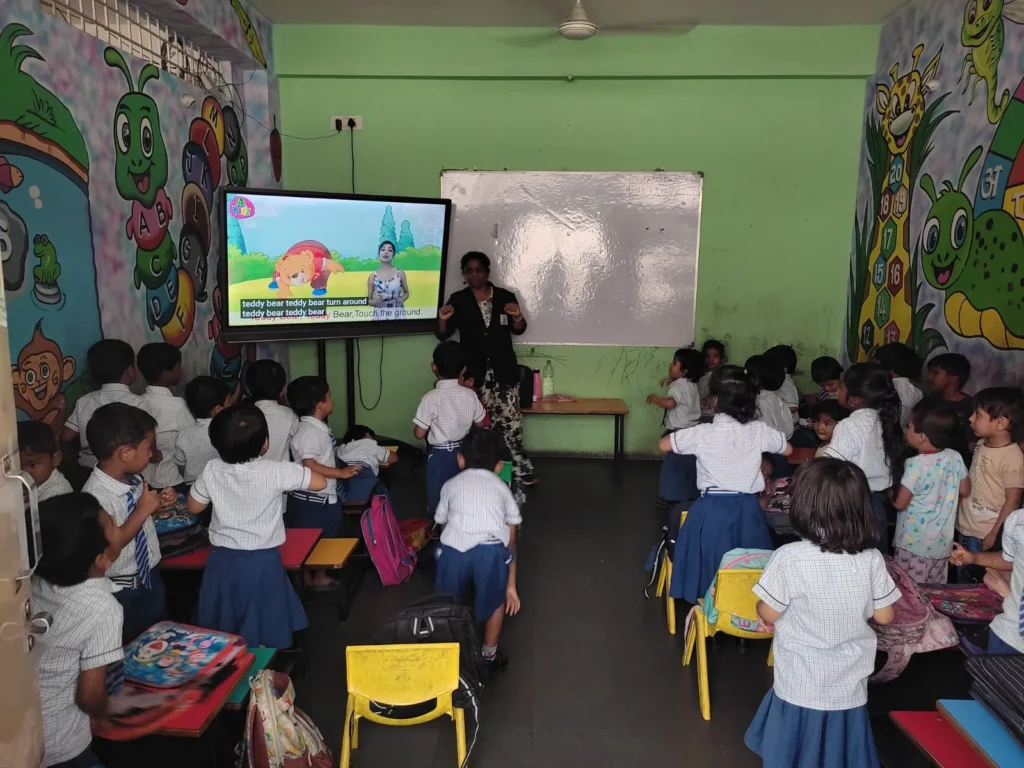 Teacher leading an interactive classroom activity while students stand near desks with digital screen and whiteboard, promoting active learning daily.