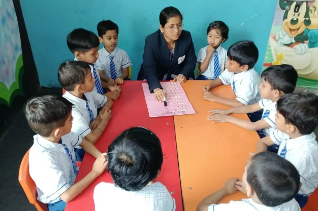 Teacher sitting with students around a table pointing at worksheet while children watch and listen during a guided group learning session.