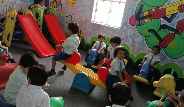 Young children in school uniform playing on slides and seesaw in an indoor play area at Gnyan Oak School with colorful wall paintings and fun learning every day.
