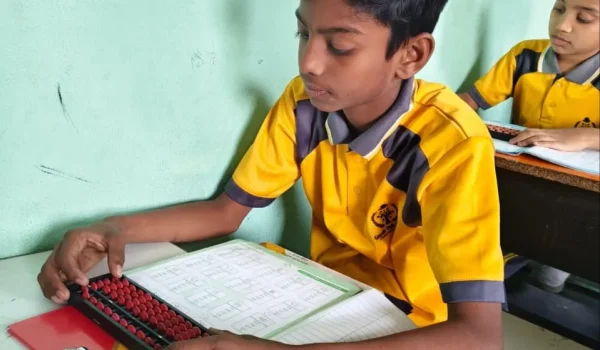 Student in yellow uniform sitting at desk writing in notebooks during classroom session with teacher nearby at Gnyan Oak School.