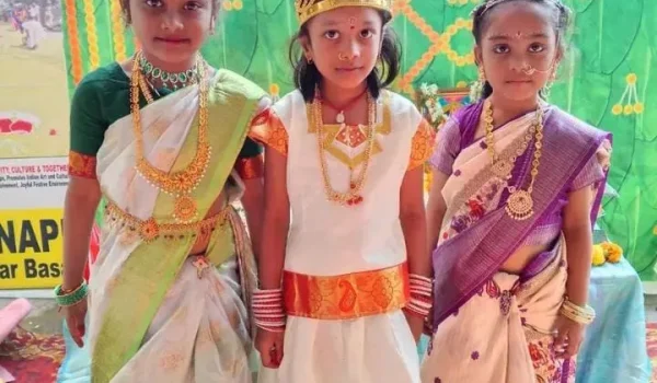 Three young girls in traditional dresses standing together wearing jewelry and colorful attire during a school cultural program.
