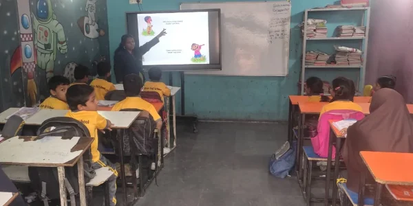 Teacher pointing at digital screen while students sit at desks inside classroom with shelves and books at Gnyan Oak School.