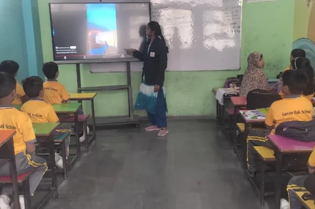 Teacher pointing at digital screen while students in yellow uniforms sit at desks inside a smart classroom at Gnyan Oak School during an interactive lesson.