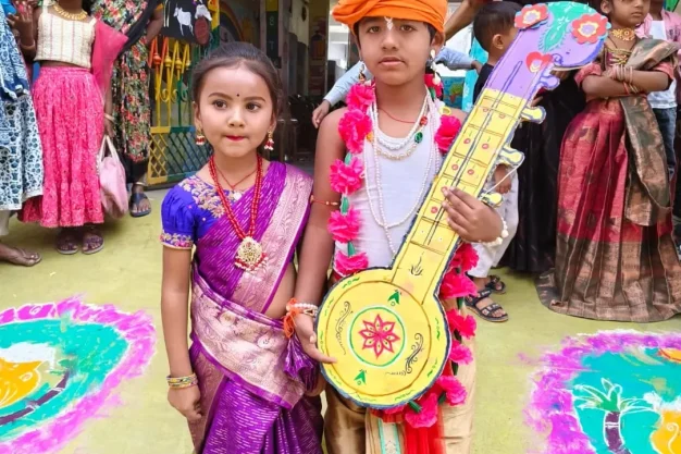 Two children in traditional attire standing with a decorative musical instrument during a colorful school cultural event.