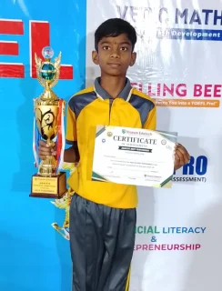 School boy in yellow uniform holding trophy and certificate in front of event backdrop at Gnyan Oak School during student achievement ceremony.